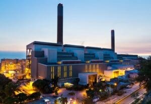 Modern industrial power plant illuminated at dusk with tall smokestacks, adjacent infrastructure, and a coastal background.